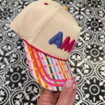 Hand holding a small beige hat with colorful brim against a patterned background