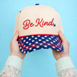 Cap with 'Be Kind' text and American flag design held against a blue background