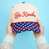 Cap with 'Be Kind' text and American flag design held against a blue background