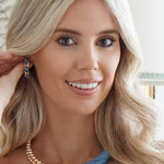Woman with blonde hair wearing a blue top and gold necklace indoors.