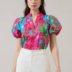 Model wearing a bright pink floral top with blue and green leaves, puff sleeves, V-neck split neckline, cropped fit, paired with white high-waisted pants
