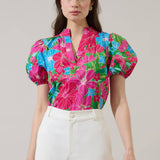 Model wearing a bright pink floral top with blue and green leaves, puff sleeves, V-neck split neckline, cropped fit, paired with white high-waisted pants
