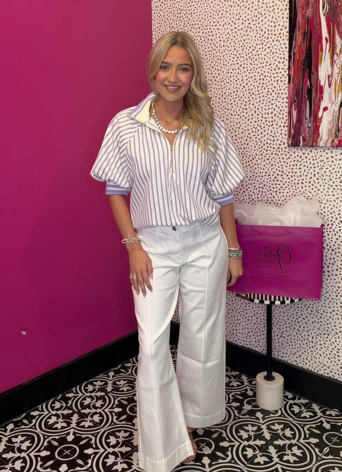 Woman in a striped shirt and white pants standing in a room with a pink wall and patterned floor.