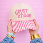 Cap with 'Uplift Others' text held by hands against a pink background